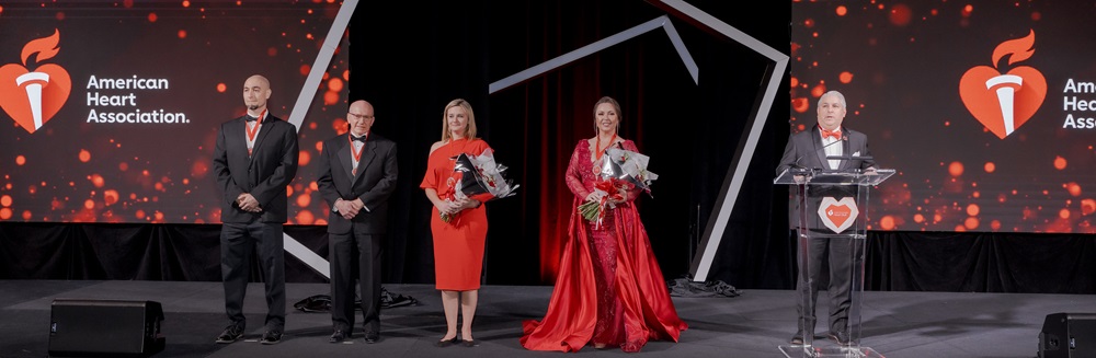Group of three men and two women in formal attire standing on stage in front of two screens projecting the American Heart Association logo.